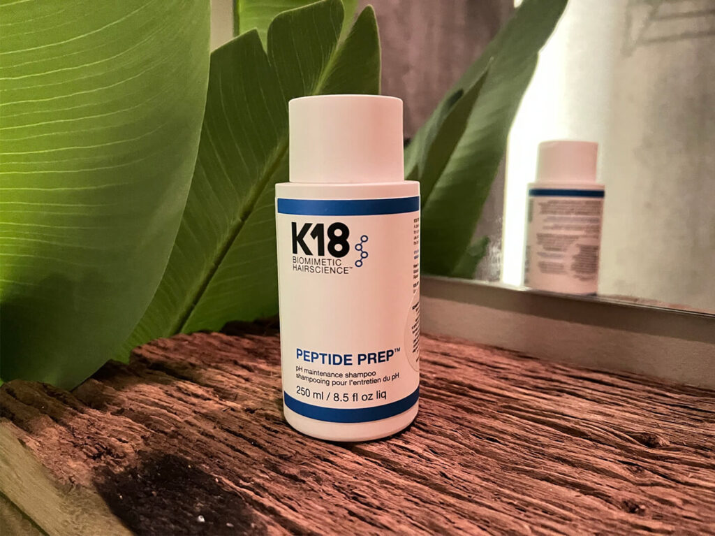 K18 Peptide Prep Shampoo stående på et træbord, med et sejl og en plante i baggrunden